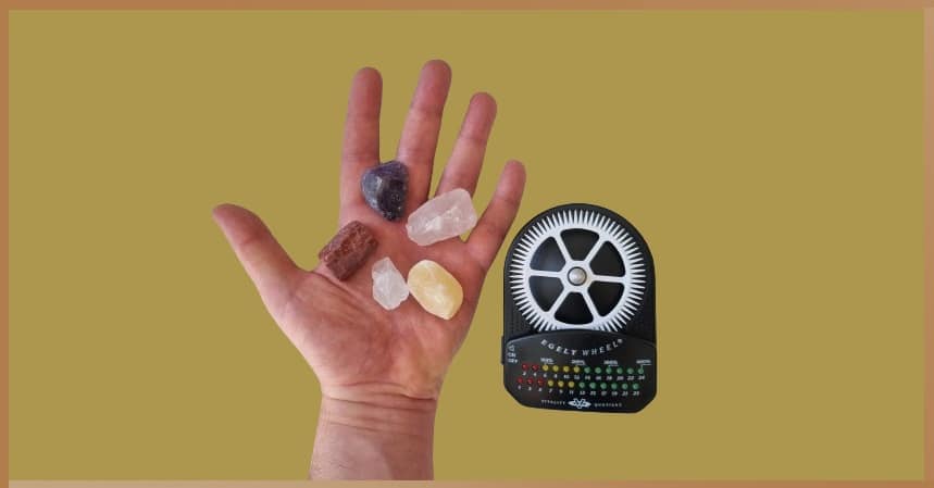 Assorted healing crystals in a hand beside an Egely Wheel device against a mustard background, symbolizing the integration of crystal energy work and bioenergy measurement for enhanced healing and balance