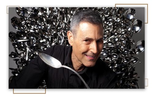 Uri Geller, a mentalist known for bending spoons, stands before a backdrop of numerous spoons, holding one as if to demonstrate his ability to manipulate objects with his mind, symbolizing the mystery of life energy.