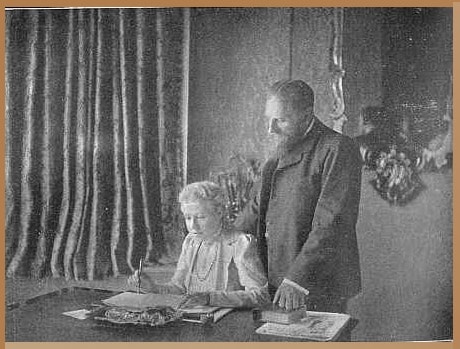 A historical black and white photograph of Annie Besant and C.W. Leadbeater in 1901, with Leadbeater standing and observing Besant, who is seated and writing at a table, surrounded by an air of concentration and mystical study.