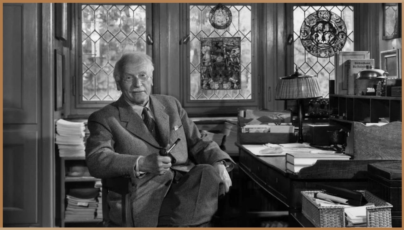 A black and white photograph of Carl Gustav Jung, seated comfortably in his study, holding a pipe, with bookshelves and a desk strewn with papers, reflective of his analytical and inquisitive nature.