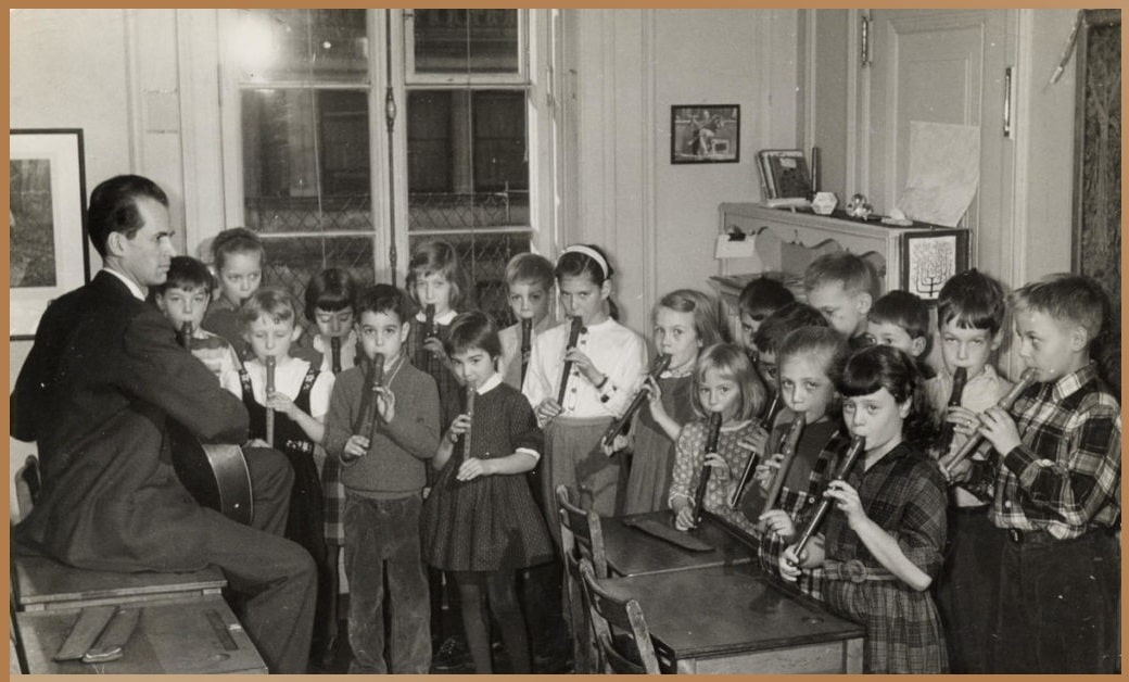 Black and white vintage photo of Rudolf Steiner teaching a group of focused children playing recorders in a classroom, exemplifying the Waldorf educational method integrating academic, artistic, and practical learning.
