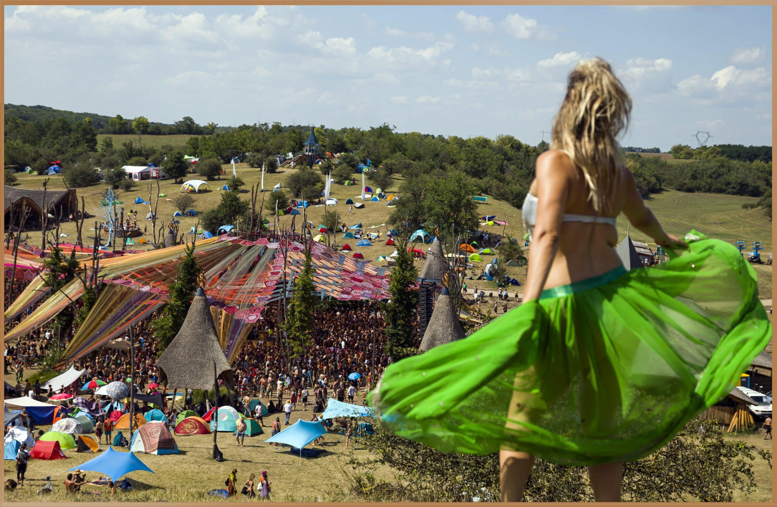 A woman in a green skirt overlooks the vibrant crowd and colorful setups at the O.Z.O.R.A. Festival in Hungary, showcasing the festival's lively atmosphere and artistic installations.