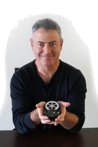 A man with salt-and-pepper hair, wearing a black V-neck sweater, holding a small device with a glowing display and intricate details.